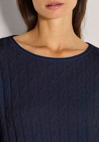 Nahaufnahme eines Frauenhalses und -brust mit einem marineblauen Kabelstrickpullover mit rundem Ausschnitt.