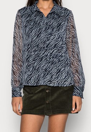 Žena v modré a černé průhledné blůze s dlouhými rukávy se zebrovým vzorem a v tmavě zelené manšestrové mini sukni na knoflíky.