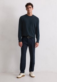 Donkerblauwe gebreide trui met een ronde hals, gecombineerd met marineblauwe broek en witte sneakers met bruine accenten. Staand voor een neutrale achtergrond.