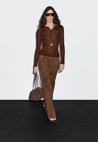 Brauner, gerippter Langarmoberteil mit V-Ausschnitt, kombiniert mit braunen Wildleder-Hosen in weiter Passform. Schwarze Sonnenbrillen und eine braune Schultertasche vollenden den Look.