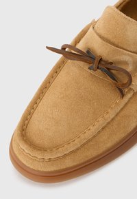 Chaussure en suede beige avec un design à bout rond, dotée d'un laçage en cuir marron et de détails cousus le long des bords. Semelle en caoutchouc fine.
