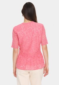 Rožinė nėrinių blusa trumpomis rankovėmis, gėlių rašto ir plačiu apvadu. Pergamentinė medžiaga, rodanti odą po ja. Nėra matomų detalumų ar akcentų.