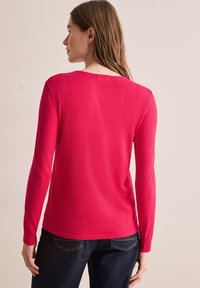 Haut rose vif à manches longues avec un col large, une coupe ajustée et des poignets côtelés, associé à un jean foncé. Texture de tissu lisse.