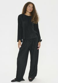 Donna con capelli ricci che indossa un top nero plissettato e pantaloni larghi, in piedi su uno sfondo bianco uniforme.