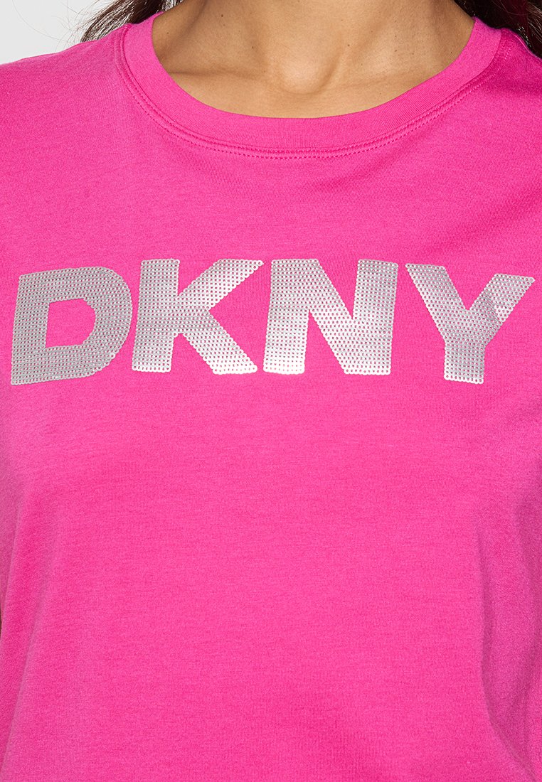 Różowy bawełnowy t-shirt z dużym srebrnym metalicznym logo "DKNY" w wyraźnej czcionce, okrągłym dekoltem i krótkimi rękawami. Gładka tekstura.