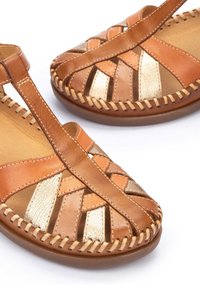 Sandali in pelle marrone con accenti dorati, design aperto, cinturini intrecciati e dettagli cuciti su una suola color sabbia. Parte superiore testurizzata.