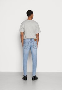 Vaqueros ajustados de color azul claro con un parche de logo en la parte trasera, combinados con una camiseta gris. Los vaqueros presentan una textura suave y detalles de costura sutiles.