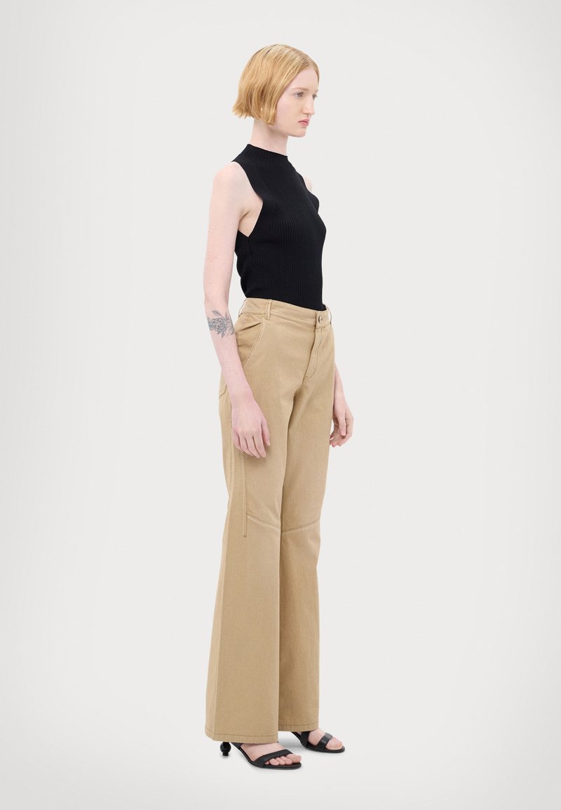 Zwarte geribbelde mouwloze top met hoge hals; beige wijde broek met een gladde textuur en zichtbare stiksels; zwarte slidesandalen.