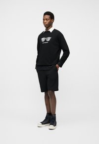 Άνδρας στέκεται με τα χέρια στις τσέπες φορώντας μαύρο φούτερ με γραφικό λευκών γυαλιών ηλίου, μαύρο σορτς και μαύρα υψηλά αθλητικά παπούτσια.