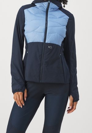 Vrouw in een marineblauwe en lichtblauwe gewatteerde zip-up jack met duimgaten en bijpassende marineblauwe legging.