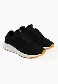 Black knit sneakers feature a textured upper, round toe, and flat laces. The sole is white with a gum rubber outsole highlighting the design.