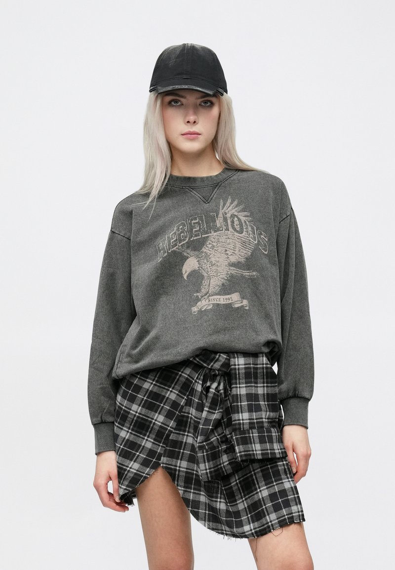 Grå hettegenser med langt erme og ørn-grafikk, kombinert med et svart- og hvitternet skjørt med knyttet midje og asymmetrisk kant.