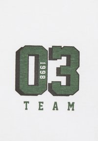 Camiseta blanca de algodón con números impresos en verde y negro "03" y "1998 TEAM" en una fuente gruesa y de bloque.