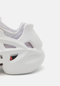 Vit sneaker med en formad design och utklipp för andningsförmåga, slät yta och ett lila nätfoder. Röda detaljer är synliga.