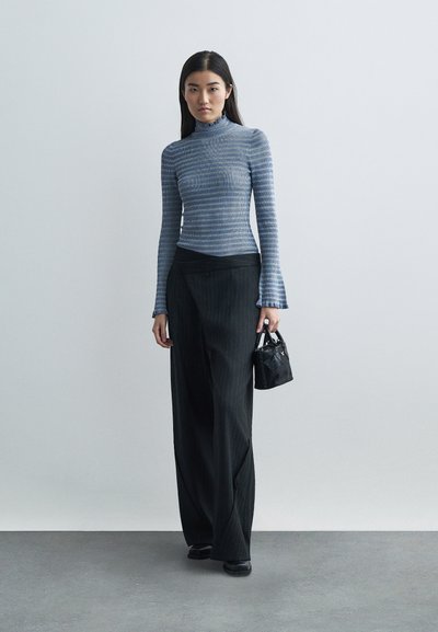 Egy nő áll, kék csíkos, hosszú ujjú felsőt, fekete csíkos, bő szárú nadrágot, fekete cipőt visel, és egy kis fekete táskával a kezében.