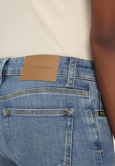 Acercamiento a unos vaqueros de denim azul con una etiqueta de cuero marrón y una pequeña etiqueta de logo, combinados con una blusa blanca de canalé.