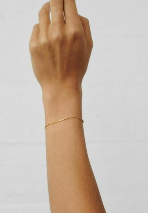 Højre arm med lysebrun hud, der bærer et delikat guldkædearmbånd mod en ensfarvet lys baggrund.