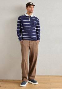Polo de mangas longas listrado em azul-marinho e branco, com colarinho branco, calças castanhas e ténis azul-marinho sobre um chão de madeira com um fundo neutro.