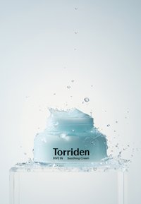 Vaaleansininen voidepurkki, jossa on teksturoitu kansi ja etiketti "Torriden DIVE IN Soothing Cream", ympäröity veden roiskeilla.