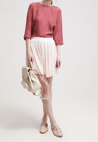 Blusa rossa a fantasia con maniche a tre quarti, gonna plissettata rosa chiaro, zaino crema e scarpe rosa pallido con dettagli a intaglio.