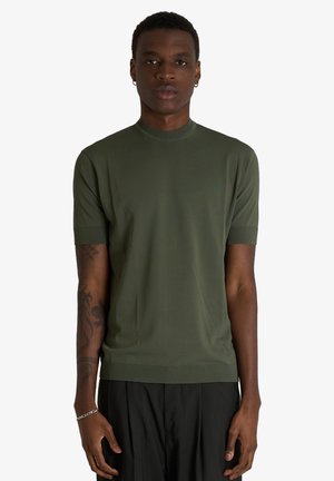 Giovane uomo dalla pelle scura che indossa una maglia verde a maniche corte lavorata a maglia e pantaloni neri, in piedi con le braccia rilassate lungo i fianchi.