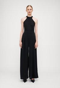 Black halter jumpsuit with wide-leg design and pleated front. Smooth fabric, high neckline, and fitted waist. Black pointed-toe shoes.