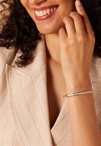 Pulsera de metal dorado con superficie lisa y una fila de cristales transparentes a lo largo de un lado. Lleva puesta en una muñeca con fondo de tela en tonos neutros.