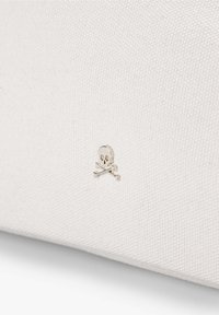Witte canvas tas met een klein zilveren doodskop en gekruiste beenderen embleem. De stof heeft een textuurafwerking, wat duurzaamheid benadrukt.
