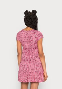 Mujer con cabello trenzado que lleva un vestido floral rojo de manga corta ajustado en la cintura, de espaldas contra un fondo claro y liso.
