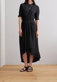 Kobieta w czarnej sukience z krótkim rękawem, zapinanej na guziki, w białe kropki, o asymetrycznym dolnym wykończeniu, w czarnych sandałach i z warstwowymi złotymi naszyjnikami.