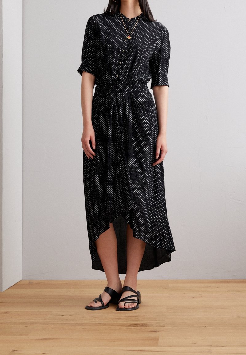 Kobieta w czarnej sukience z krótkim rękawem, zapinanej na guziki, w białe kropki, o asymetrycznym dolnym wykończeniu, w czarnych sandałach i z warstwowymi złotymi naszyjnikami.
