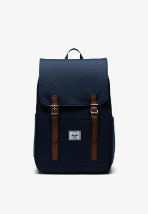 Granatowy plecak z płótna z brązowymi skórzanymi paskami i metalowymi klamrami, z zasuwaną górną klapą, przednią kieszenią oraz małą etykietą marki.