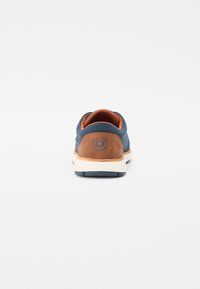 Zapatilla de tela azul marino con acentos de cuero marrón, entresuela blanca y parte trasera texturizada. Presenta un logotipo circular en el talón.