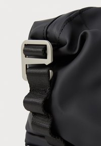 Fekete nylon táska sarka texturált felülettel, fényes ezüst színű fém csattal és állítható szövött pánttal.
