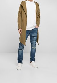 Larga chaqueta de punto verde olivo con capucha sobre una camiseta blanca, combinada con jeans azules desgastados y zapatillas blancas. Diseño sencillo y corte relajado.