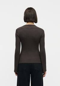 Γυναίκα με κοντά καστανά μαλλιά φοράει στενό σκούρο καφέ μακρυμάνικο τοπ και μαύρο παντελόνι, στραμμένη με την πλάτη προς ένα απλό λευκό φόντο.