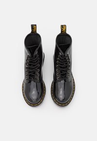 Ett par svarta blanka läder kängor med snörning och gula sömmar, med dragremmar, sett ovanifrån på en vit bakgrund.