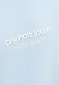 Tessuto blu chiaro con una texture liscia che presenta testo in rilievo bianco: "Originals Studio" e "portato a voi da JACK&JONES."