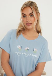 Vaaleansininen puuvillat-paita lyhyillä hihoilla, jossa on kukkakuvio ja teksti "nauti jokaisesta hetkestä" valkoisella käsinkirjoituksella.