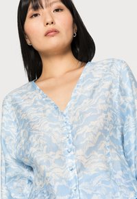 Blusa azul clara com decote em V, fecho de botão e um padrão abstrato texturizado em branco. Mangas longas com um corte descontraído.