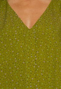 Tela verde con escote en V, que presenta un pequeño patrón de lunares negros y acentos florales dispersos en morado y verde.
