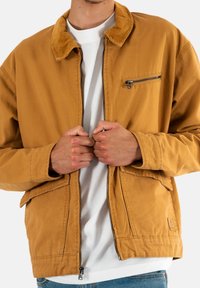 Giacca di cotone marrone con colletto in velluto a coste, zip frontale e tasca sul petto. Presenta tasche a toppa e una vestibilità rilassata, indossata sopra una camicia bianca.