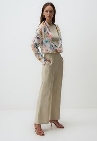 Blusa trasparente a fantasia floreale in colori pastello, indossata sopra un top fitted beige. Pantaloni palazzo beige a vita alta e tacchi nudi.