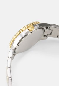 Montre en acier inoxydable avec un cadran doré et une couronne, finition polie, maillons intégrés, et un bord texturé sur le cadran.