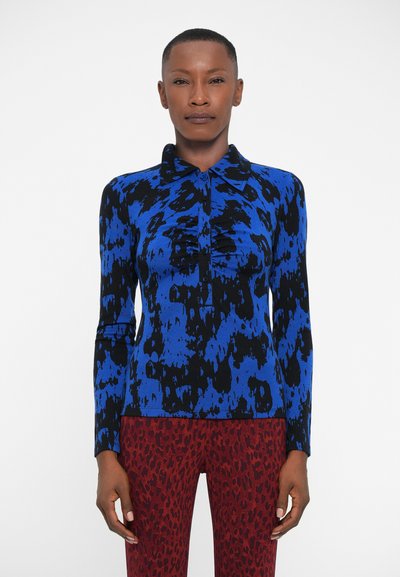 Blauw en zwart langomshirt met een kraag, voorzien van een gestructureerde ruffle langs de knopenlijst, gecombineerd met rode broek met luipaardprint.