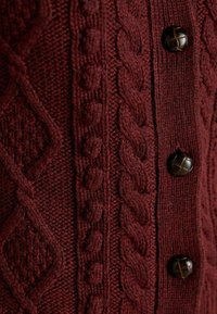 Burgundská pletená cardigan s reliéfnými kabelkovými vzormi, hladkými akcentmi a tmavo hnedými gombíkmi, čím vytvára bohatý, vrstvený vzhľad.