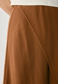 Hnědá látková sukně s jemnou texturou, šikmým švem a skládající se designem, který elegantně splývá.