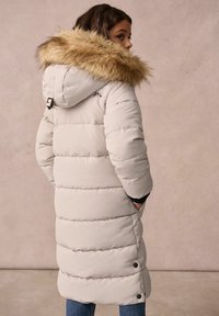 Hellbeige Daunenjacke mit einer mit Fell besetzten Kapuze, horizontal gesteppten Einsätzen, knielangem Schnitt und seitlichen Knopfverschlüssen.