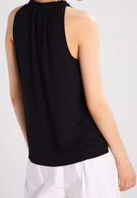 Svart ärmlös topp med en samlad halsringning, texturerat tyg och rundad nederkant, i kombination med ljusa byxor.