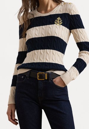 Vrouw draagt een kabelgebreide trui met brede marineblauwe en beige strepen, gouden ankerembleem op de borst, donkere spijkerbroek en zwarte riem met gouden gesp.
